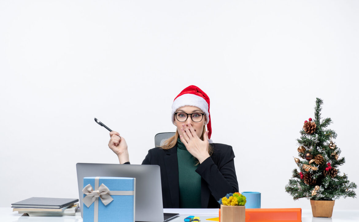 surprised-blonde-woman-with-santa-claus-hat-sitting-table-with-christmas-tree-gift-it-white-background