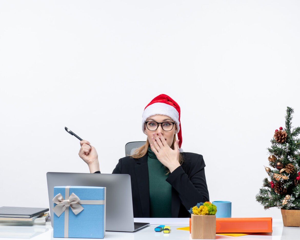 surprised-blonde-woman-with-santa-claus-hat-sitting-table-with-christmas-tree-gift-it-white-background